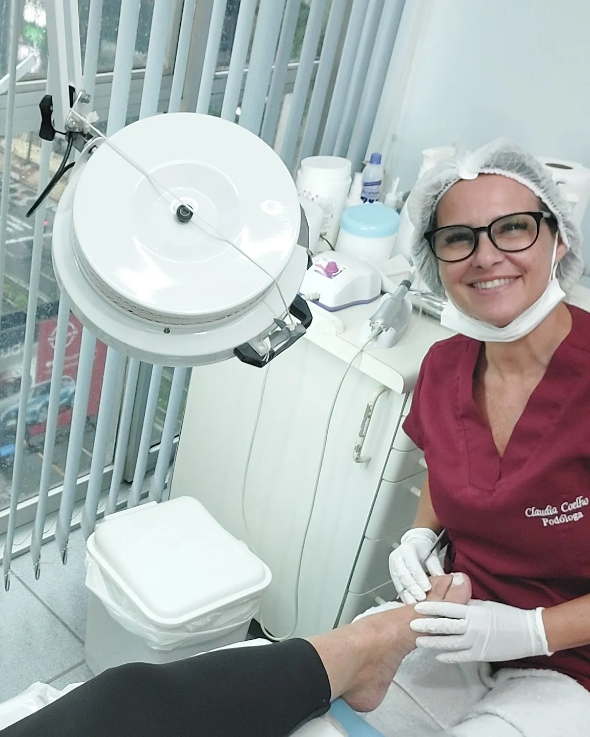 Sala de podologia com janela para a Avenida Ana Costa, foco de luz e a podóloga atendendo ao fundo