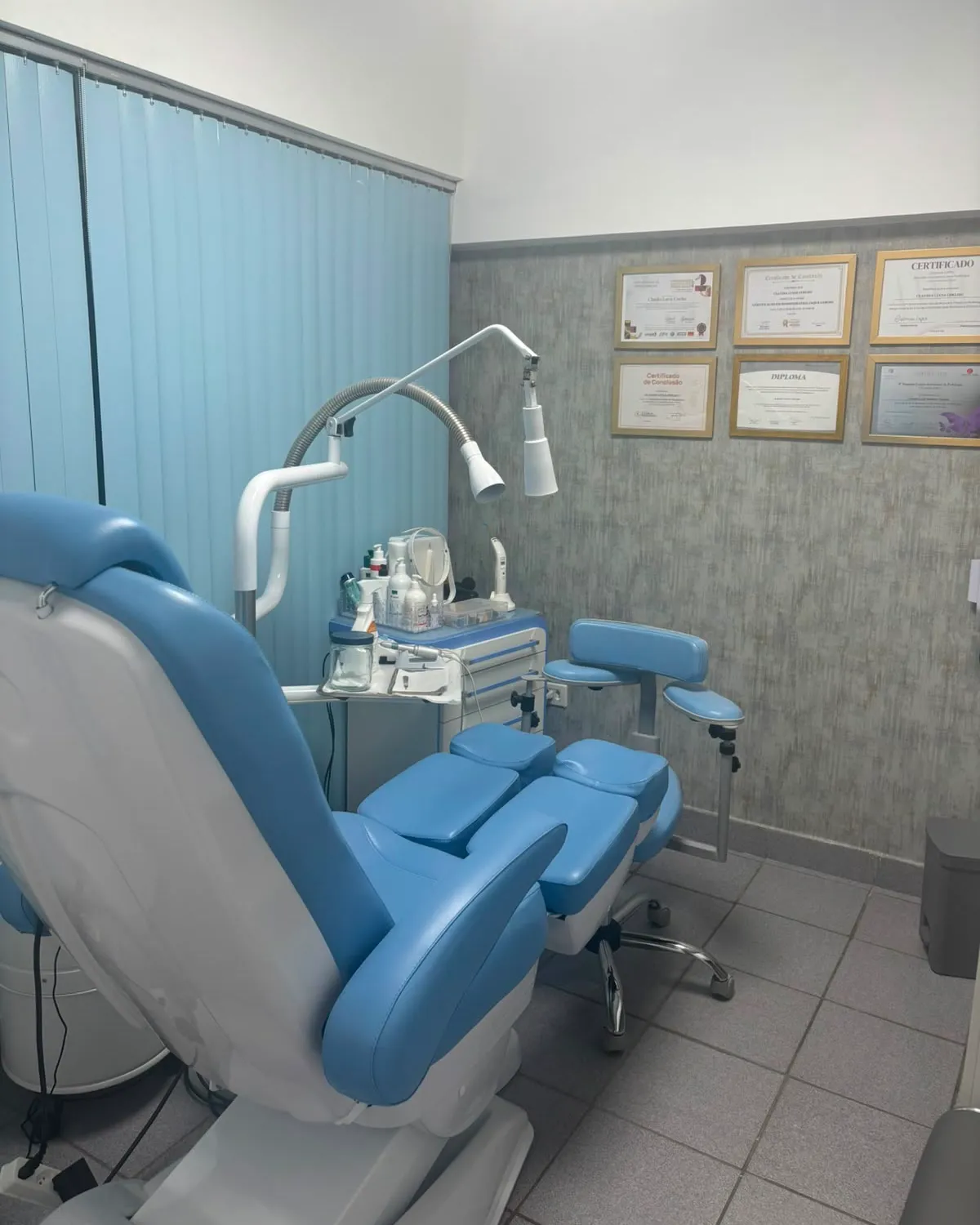 Sala de podologia com cadeira azul reclinável, foco de luz, carrinho de instrumentos e certificados emoldurados na parede