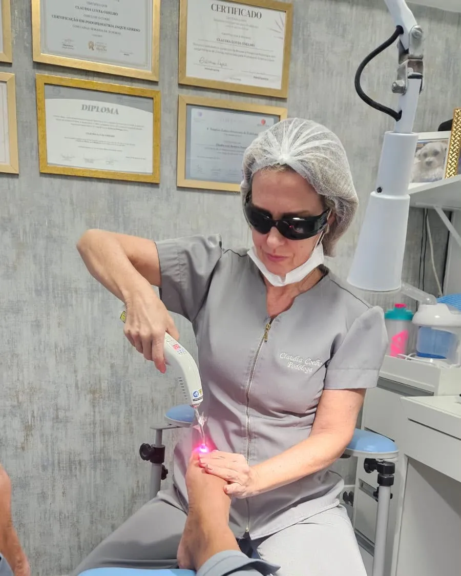 Claudia aplicando o laser em um pé, de óculos de proteção e touca, com a parede de certificados ao fundo