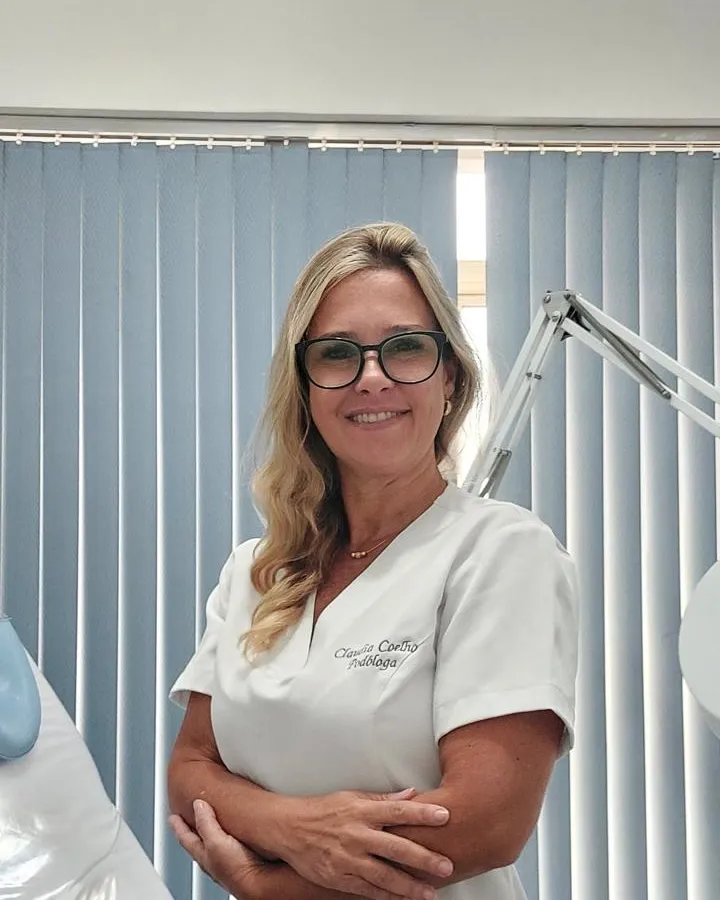 Claudia Coelho de jaleco branco bordado com o nome dela, na sala de atendimento, com as persianas azuis ao fundo